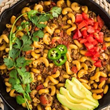A skillet filled with macaroni, ground meat, and sauce, topped with sliced avocado, chopped tomatoes, jalapeño, and cilantro.