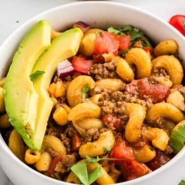 A bowl of macaroni with ground beef, tomato, herbs, and avocado slices, with cherry tomatoes and cilantro on the side.