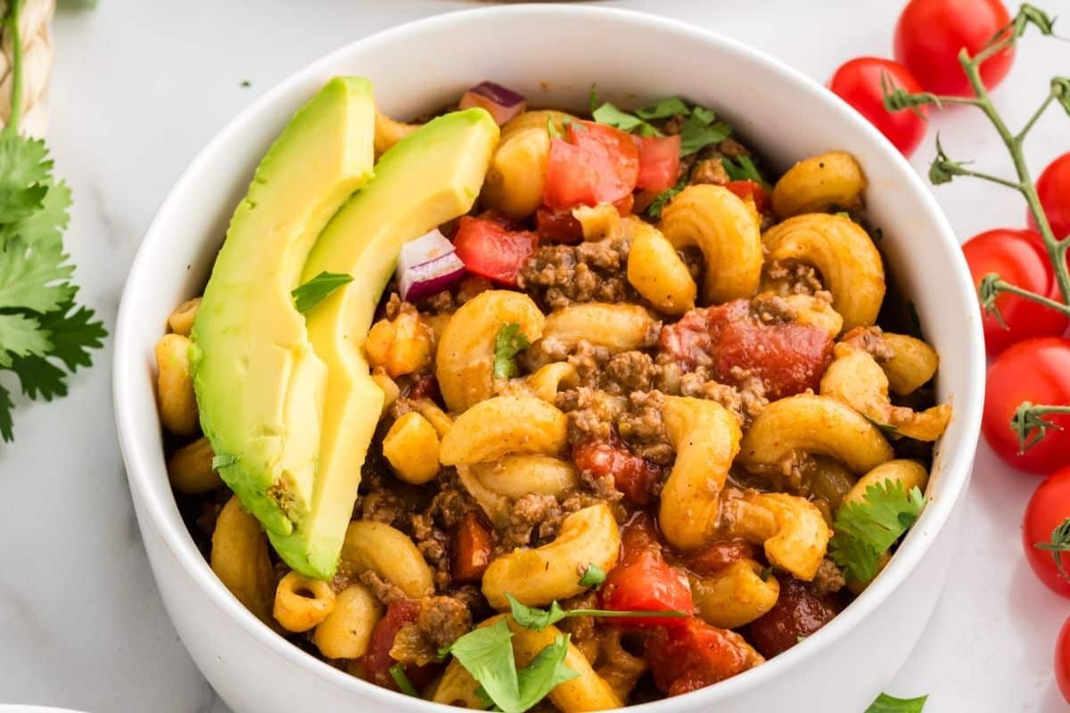 A bowl of macaroni with ground beef, tomato, herbs, and avocado slices, with cherry tomatoes and cilantro on the side.