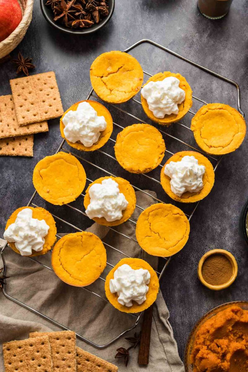 Mini pumpkin cheesecake bites on a cooling rack, some topped with whipped cream, surrounded by graham crackers, spices, and baking ingredients on a dark surface.