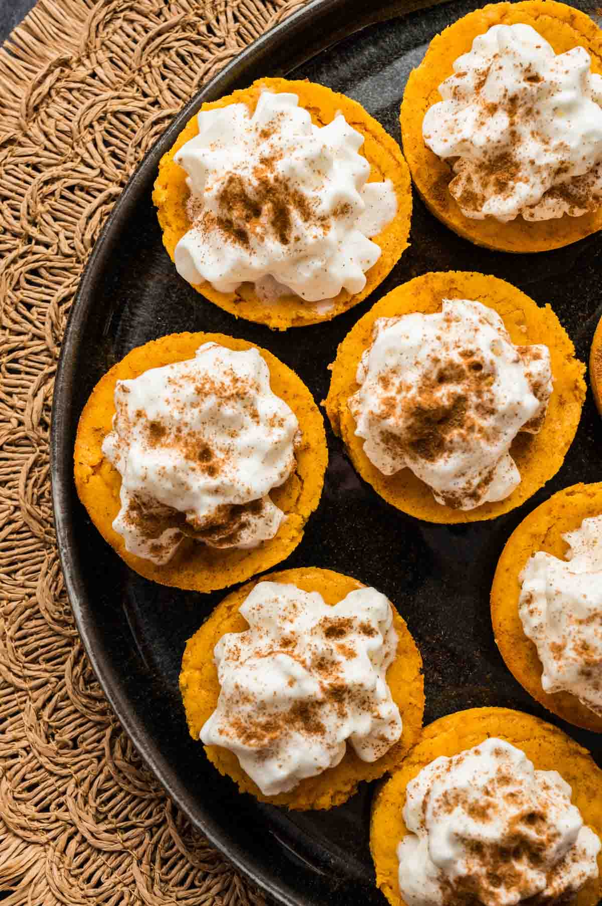 A black plate with several pumpkin cheesecake bites topped with whipped cream and sprinkled with cinnamon, placed on a woven mat.