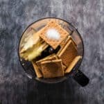 A food processor bowl filled with graham crackers, butter, and sugar, viewed from above on a dark countertop.