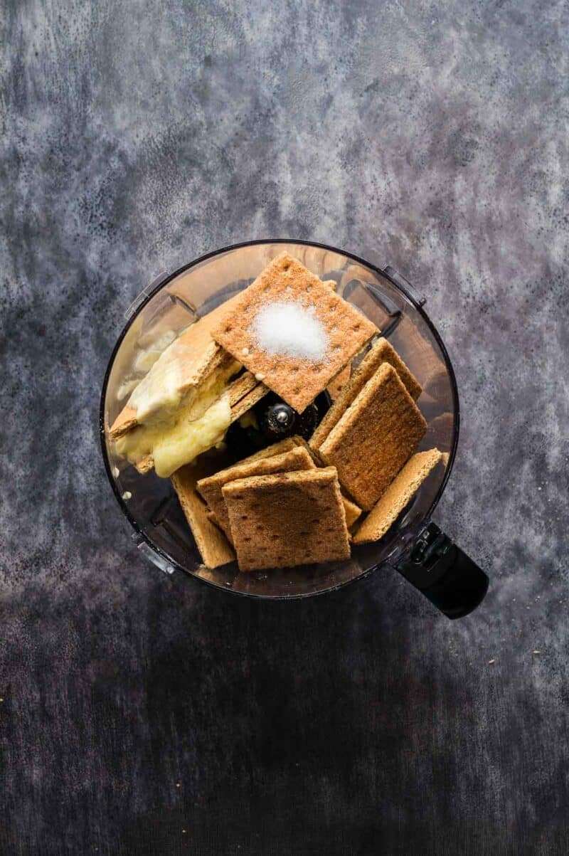 A food processor bowl filled with graham crackers, butter, and sugar, viewed from above on a dark countertop.