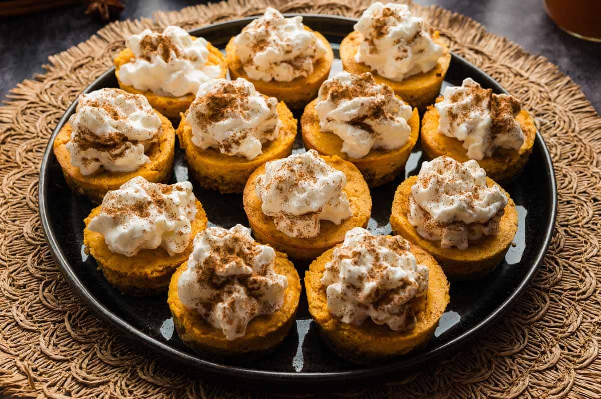 A plate of small pumpkin cheesecake bites topped with whipped cream and sprinkled with cinnamon, arranged in rows on a black plate.