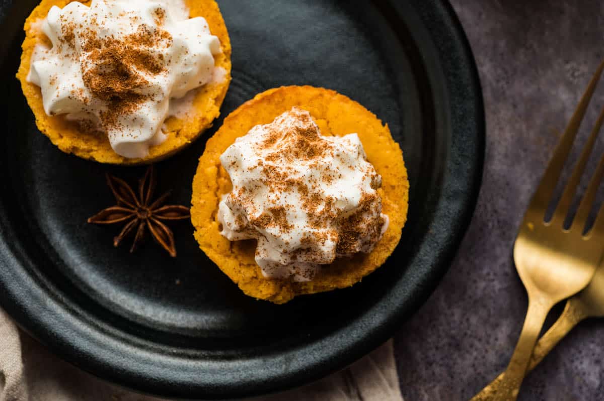 Two mini pumpkin cheesecake bites topped with whipped cream and sprinkled with cinnamon, served on a black plate with a star anise. A gold fork is placed to the side.