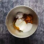 A mixing bowl containing pumpkin puree, sugar, egg, yogurt, baking powder, and baking soda on a dark countertop.