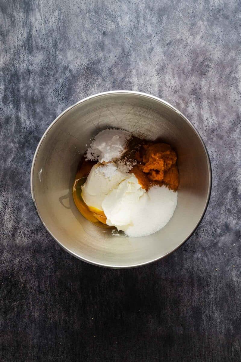 A mixing bowl containing pumpkin puree, sugar, egg, yogurt, baking powder, and baking soda on a dark countertop.