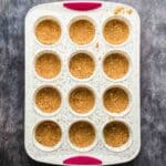 A muffin tin with twelve cups, each filled with a pressed crumb crust, sits on a dark textured surface.