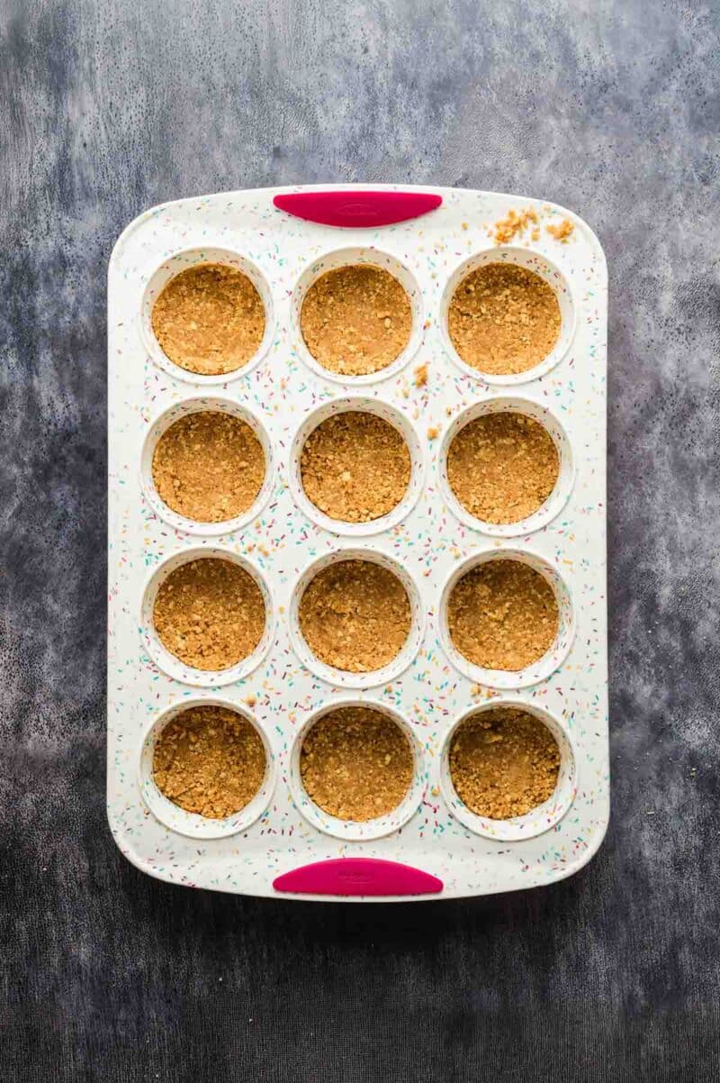 A muffin tin with twelve cups, each filled with a pressed crumb crust, sits on a dark textured surface.