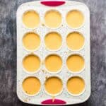A 12-cup muffin tin filled with yellow batter sits on a dark surface. The tin has colorful speckles and two red silicone grips.