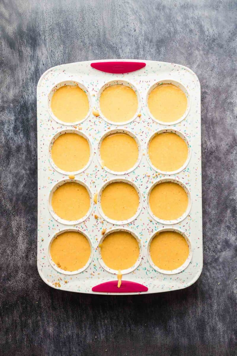 A 12-cup muffin tin filled with yellow batter sits on a dark surface. The tin has colorful speckles and two red silicone grips.