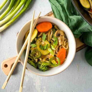 A bowl of vegetable stir-fry noodles inspired by Japanese cuisine, with carrots, zucchini, mushrooms, broccoli, and green onions, garnished with sliced jalapeños. Two chopsticks rest on the bowl.