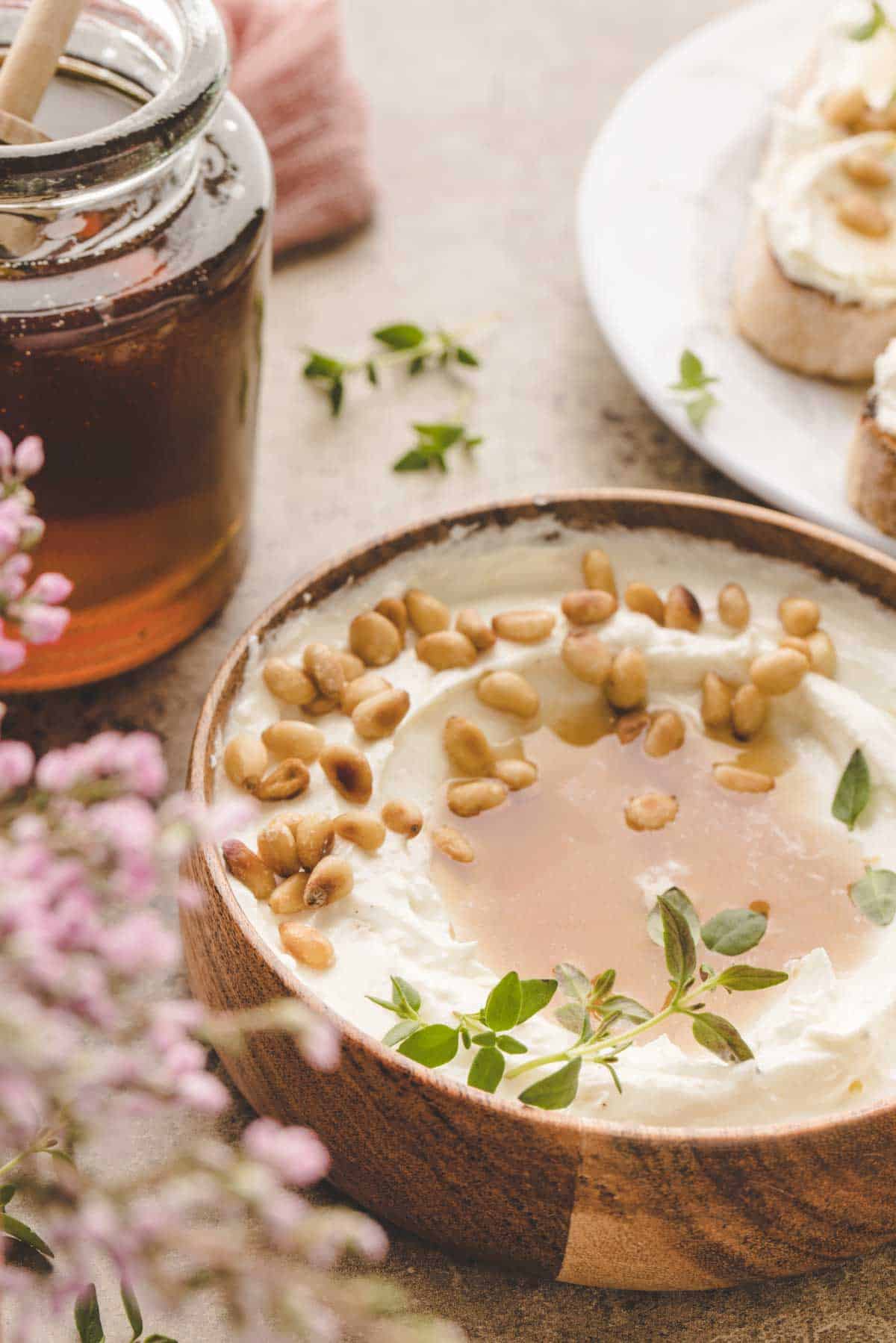 A wooden bowl of whipped ricotta topped with honey, toasted pine nuts, and fresh herbs sits next to a jar of honey and a plate of crostini.