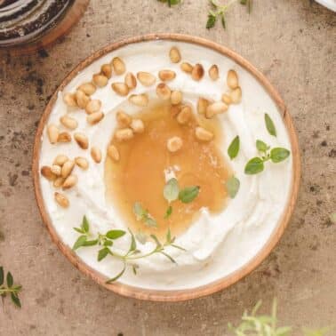 A wooden bowl of whipped ricotta topped with honey, pine nuts, and fresh thyme on a stone surface, with herb sprigs scattered nearby.
