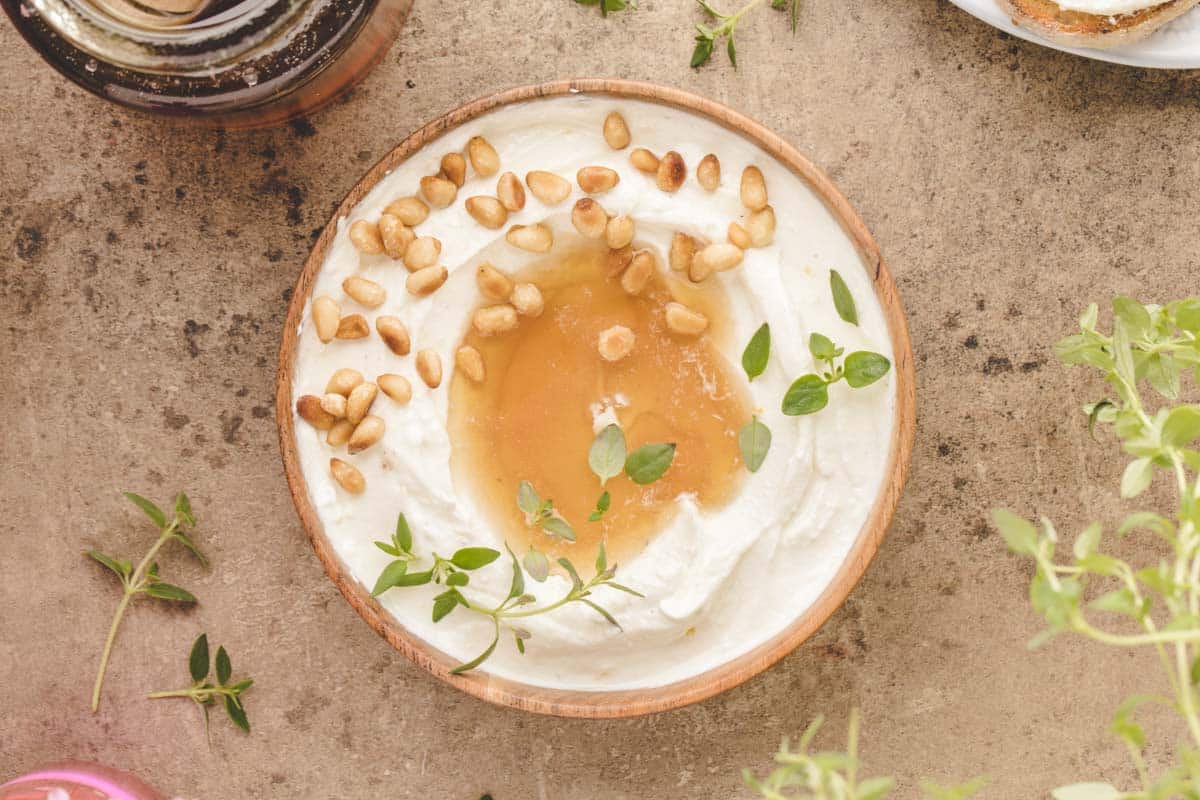 A wooden bowl of whipped ricotta topped with honey, pine nuts, and fresh thyme on a stone surface, with herb sprigs scattered nearby.