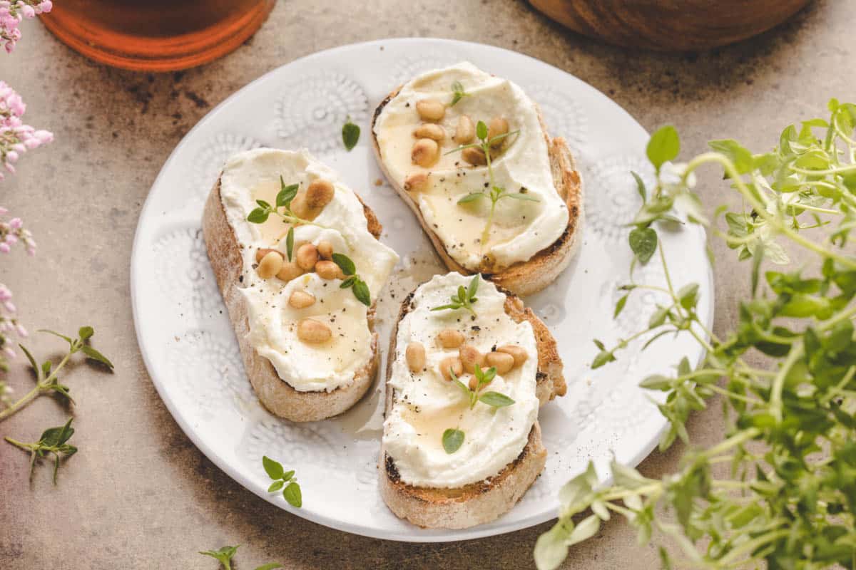 Three slices of bread with creamy cheese, pine nuts, honey, and fresh herbs are arranged on a white plate.