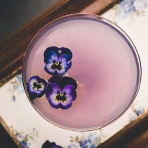 A pink cocktail garnished with three purple pansy flowers, viewed from above, on a floral-patterned tray.