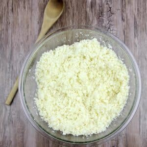 A glass bowl filled with cauliflower rice on a wooden surface, with a wooden spoon placed beside the bowl.