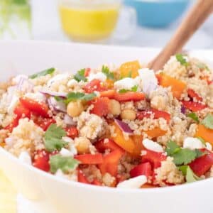 A bowl of quinoa salad with chickpeas, red bell peppers, red onions, feta cheese, and cilantro, with a wooden spoon for serving.