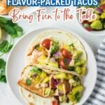 Two soft shell, flavor-packed tacos filled with grilled fish, diced tomatoes, onions, avocado, and sauce on a white plate, garnished with cilantro and more food visible in the background.