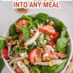 A bowl of bright greens spinach salad topped with shredded chicken, sliced strawberries, walnuts, and cheese crumbles, placed on a light surface for a fresh, savory flavor.