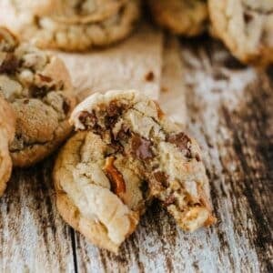 A chocolate chip cookie broken in half sits on a rustic wooden surface, with more cookies and a piece of parchment paper in the background.