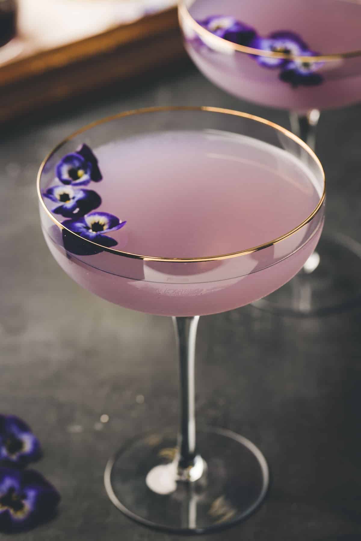 A coupe glass filled with a light purple cocktail, garnished with purple edible flowers, sits on a dark surface. Another similar glass is partially visible in the background.