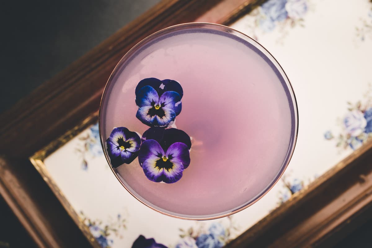 A pink cocktail in a glass, garnished with three purple and white edible flowers, viewed from above on a floral-patterned tray.