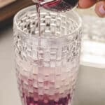 Purple liquid is being poured from a small glass pitcher into a textured glass filled with ice and a light purple beverage.