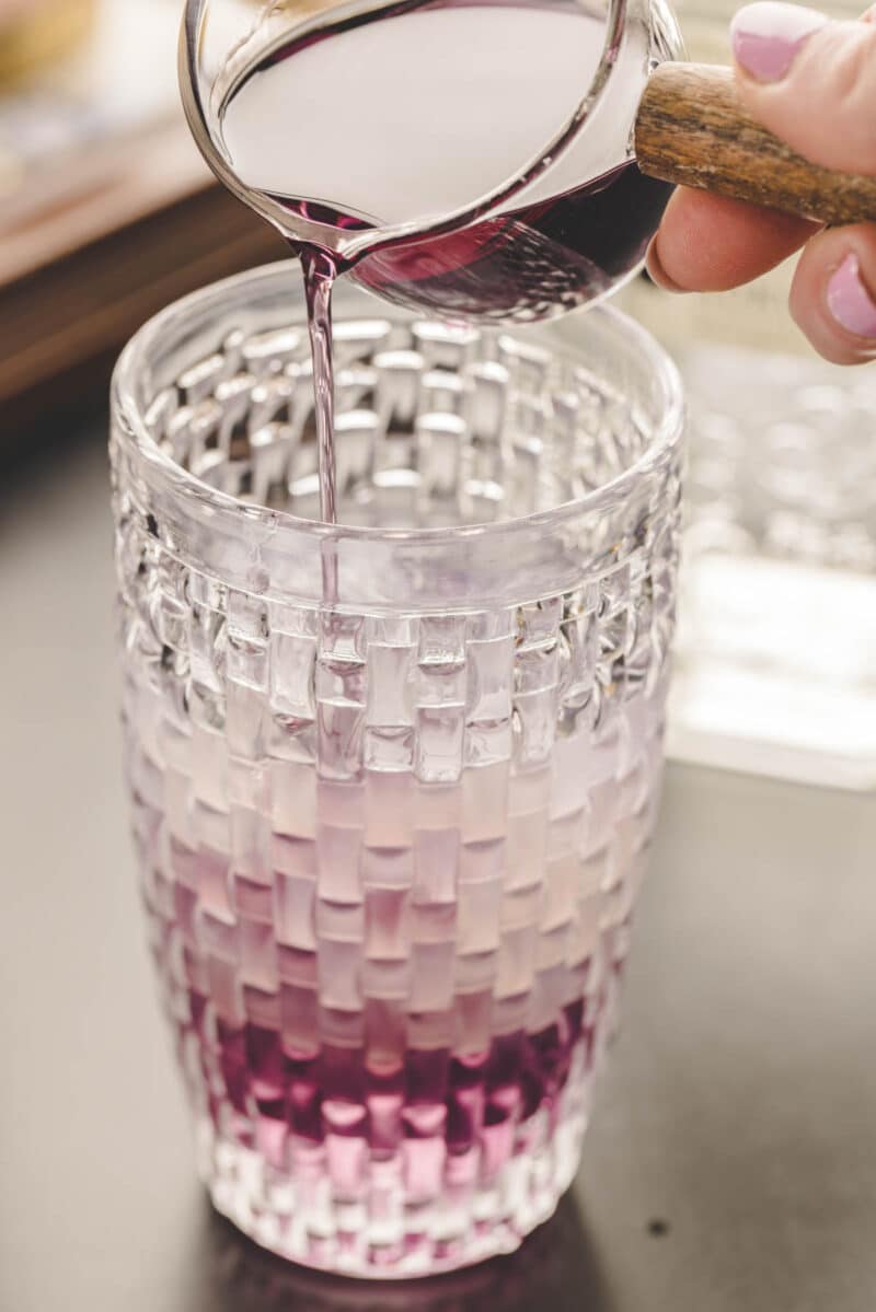 Purple liquid is being poured from a small glass pitcher into a textured glass filled with ice and a light purple beverage.