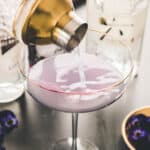 A cocktail shaker pours a light purple drink into a stemmed glass, with bottles and purple flowers nearby on a tabletop.