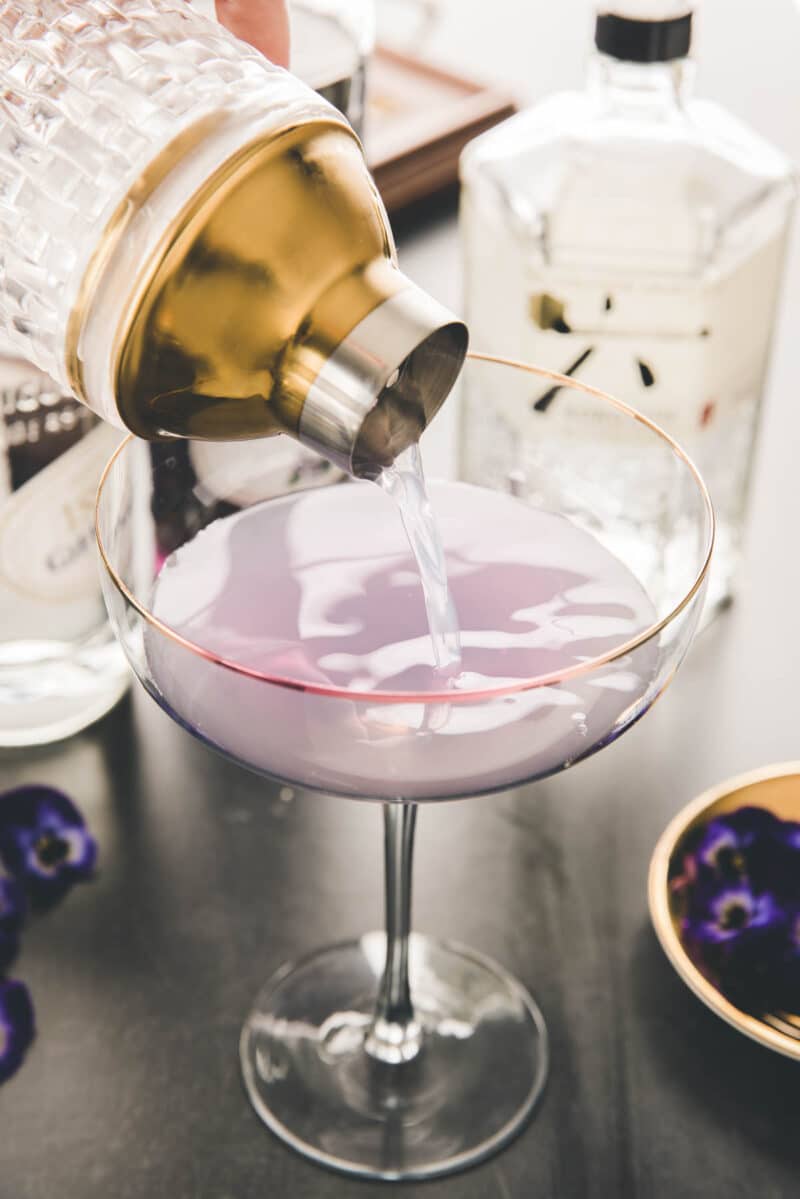 A cocktail shaker pours a light purple drink into a stemmed glass, with bottles and purple flowers nearby on a tabletop.