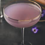 A coupe glass filled with a pale purple cocktail, with a gold rim on the glass, set on a dark surface with small purple flowers nearby.