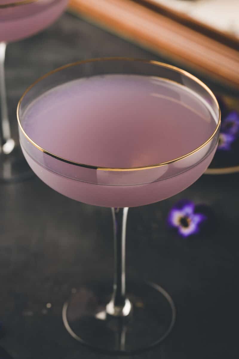 A coupe glass filled with a pale purple cocktail, with a gold rim on the glass, set on a dark surface with small purple flowers nearby.