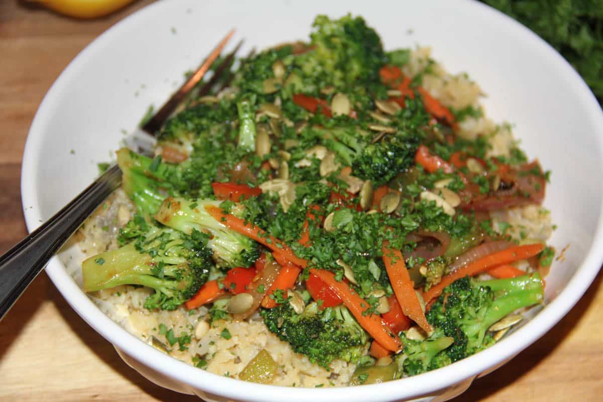 A bowl of sautéed broccoli, carrots, and onions served over rice or grains, garnished with chopped herbs and seeds, with a fork on the side.