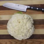 A whole cauliflower sits on a wooden cutting board next to a kitchen knife.