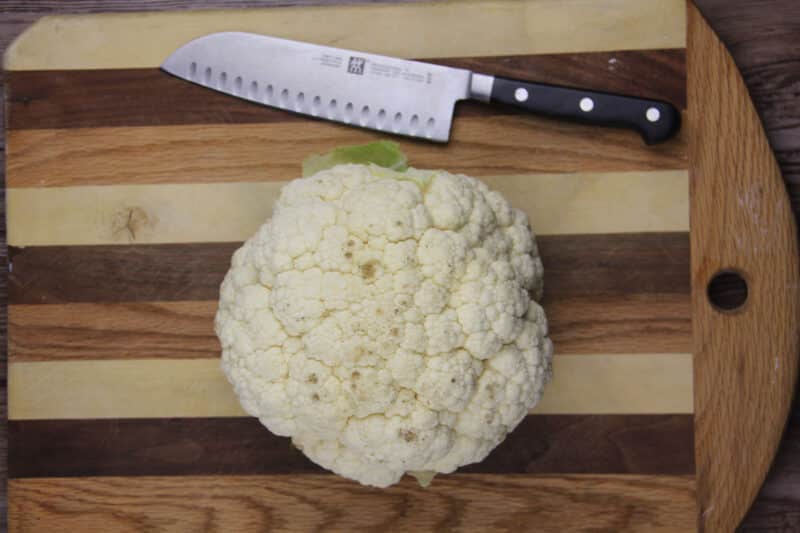 A whole cauliflower sits on a wooden cutting board next to a kitchen knife.