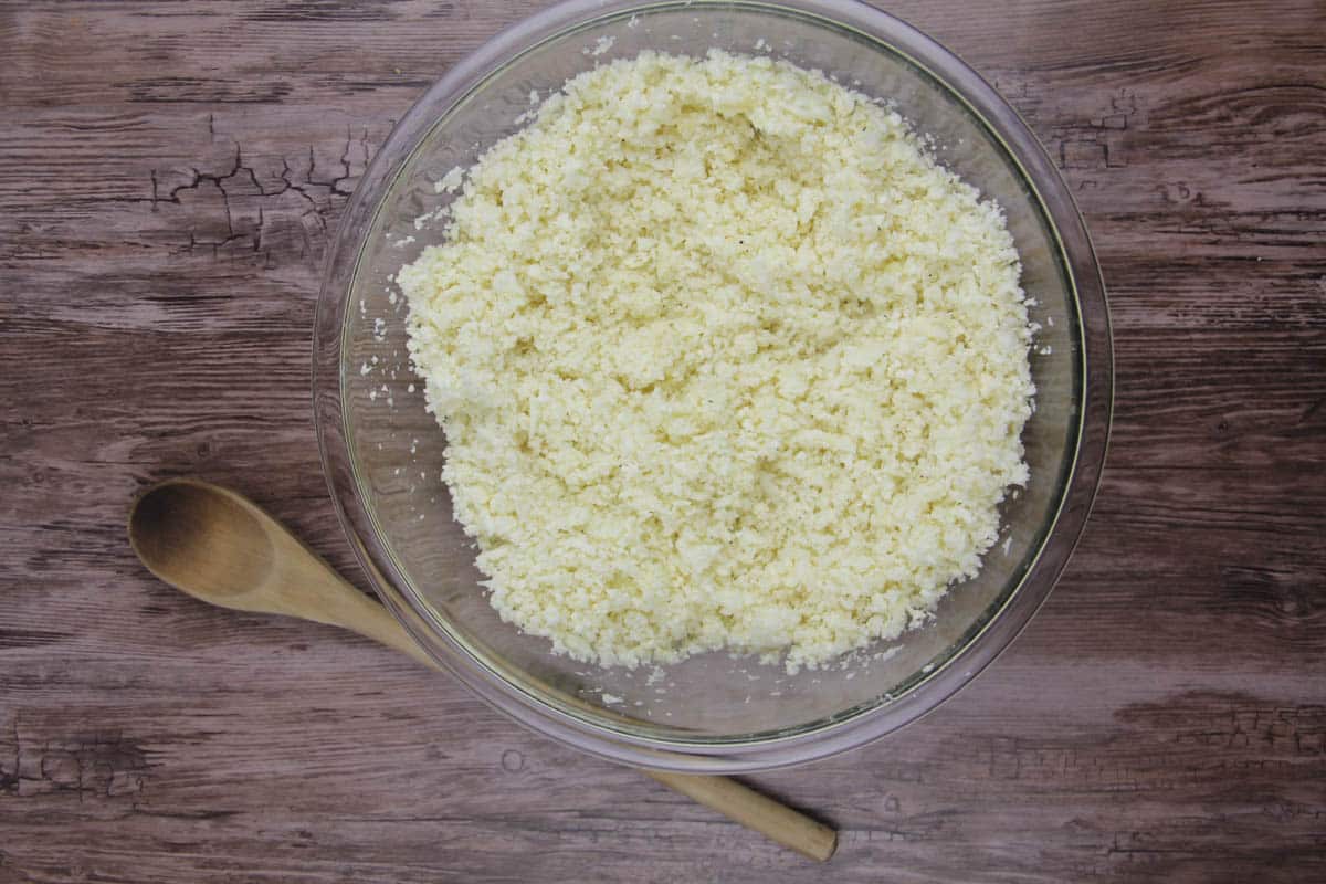 A glass bowl filled with finely riced cauliflower sits on a wooden surface next to a wooden spoon.