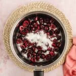 A stainless steel saucepan with halved cherries and a sprinkling of sugar inside, placed on a woven mat next to a pink cloth on a light pink surface.