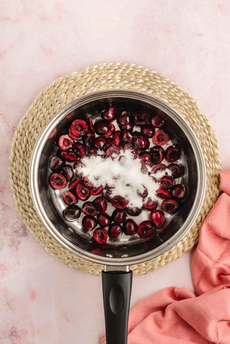 A stainless steel saucepan with halved cherries and a sprinkling of sugar inside, placed on a woven mat next to a pink cloth on a light pink surface.