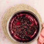 A saucepan containing cooked cherries in syrup rests on a woven mat, with a pink cloth partially visible nearby.
