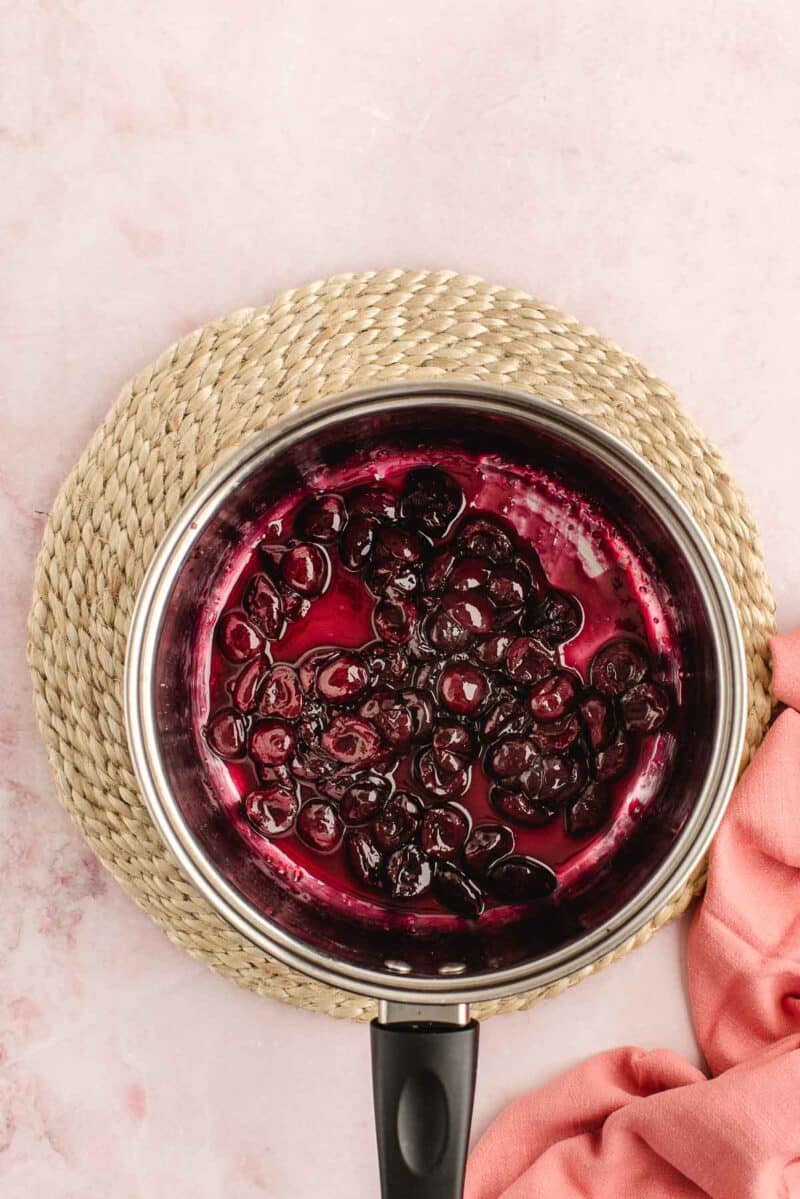 A saucepan containing cooked cherries in syrup rests on a woven mat, with a pink cloth partially visible nearby.