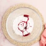 A glass bowl containing powdered sugar and red liquid sits on a woven placemat, with a pink cloth partially visible nearby.