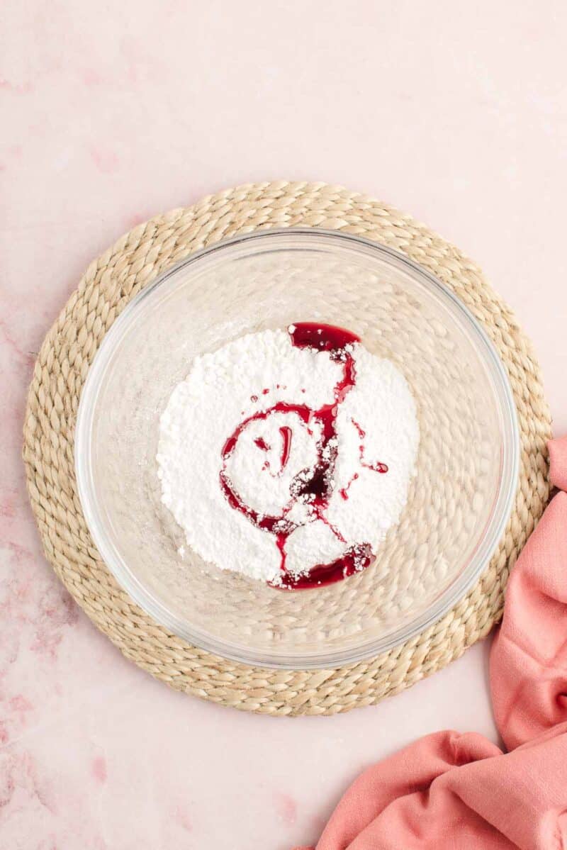 A glass bowl containing powdered sugar and red liquid sits on a woven placemat, with a pink cloth partially visible nearby.