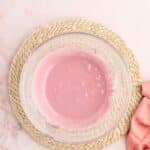 A clear glass bowl filled with pink liquid sits on a woven mat, with a pink cloth partially visible to the side.