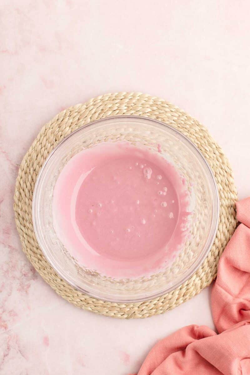 A clear glass bowl filled with pink liquid sits on a woven mat, with a pink cloth partially visible to the side.