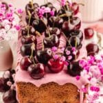 A rectangular loaf cake with pink icing, topped with fresh cherries and decorated with small pink flowers, sits on a white surface.