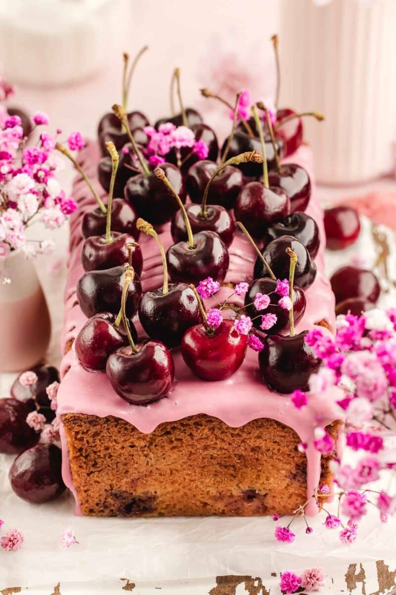 A rectangular loaf cake with pink icing, topped with fresh cherries and decorated with small pink flowers, sits on a white surface.