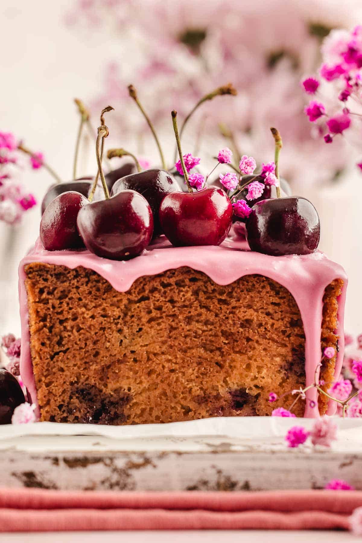 A loaf cake with pink icing, topped with fresh cherries and small pink flowers, is displayed with a soft-focus floral background.