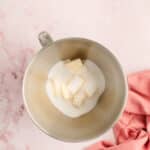 A metal mixing bowl with sugar and cubed butter inside, placed on a pink surface next to a pink cloth.